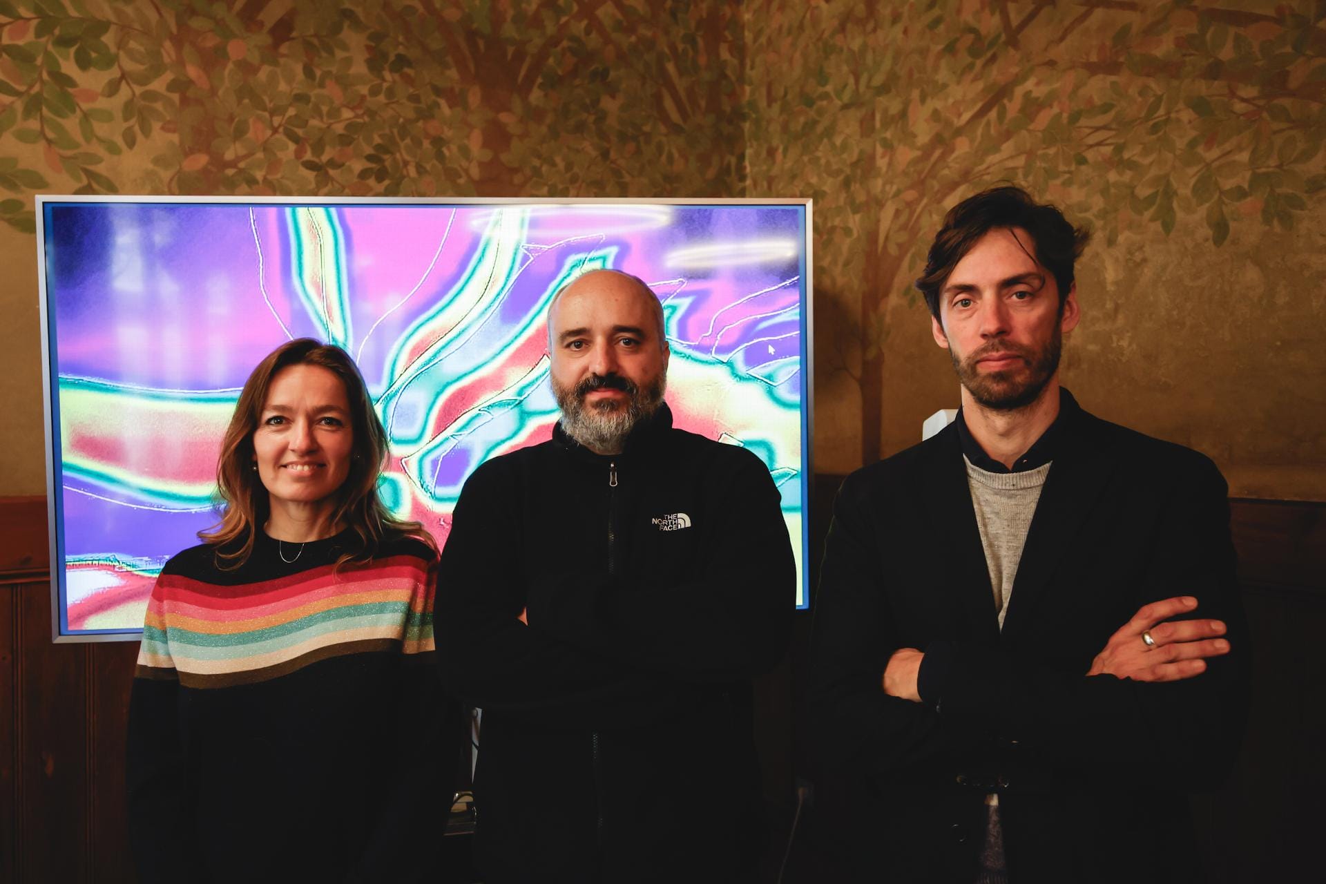 A woman in a black sweater with colorful stripes, a bald man in a black sports jacket, and a man in a black suit jacket stand in front of a screen with abstract video art.