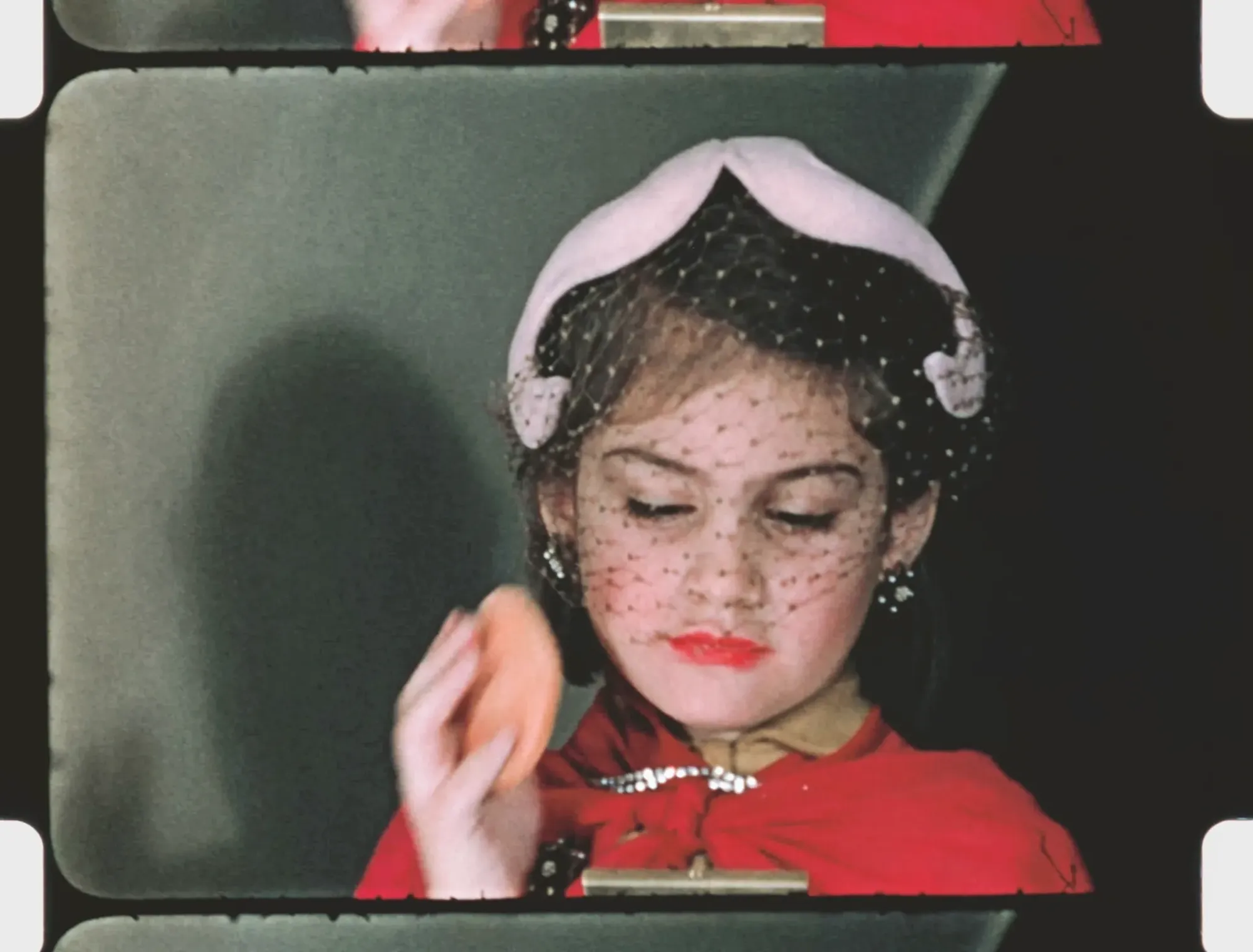 A film frame with exposed sprocket holes, with a young woman wearing a white cap and veil putting makeup on herself.