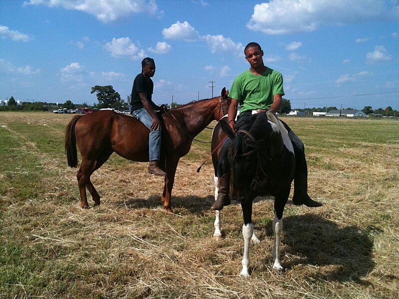 Two young Black men sitting on horses in an open field with a blue sky behind them.