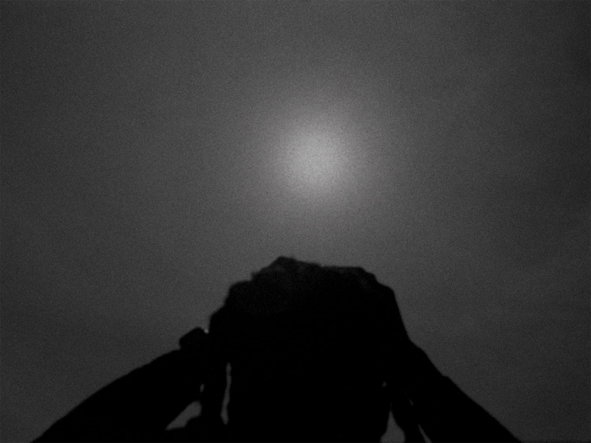A grainy black and white image of a figure in shadows holding binoculars up to their face, with a hazy sun in the background.
