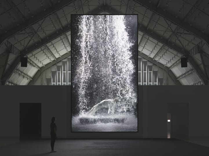 A large, vertical video screen hanging in a dark gallery with an image of a figure with water raining down on them.