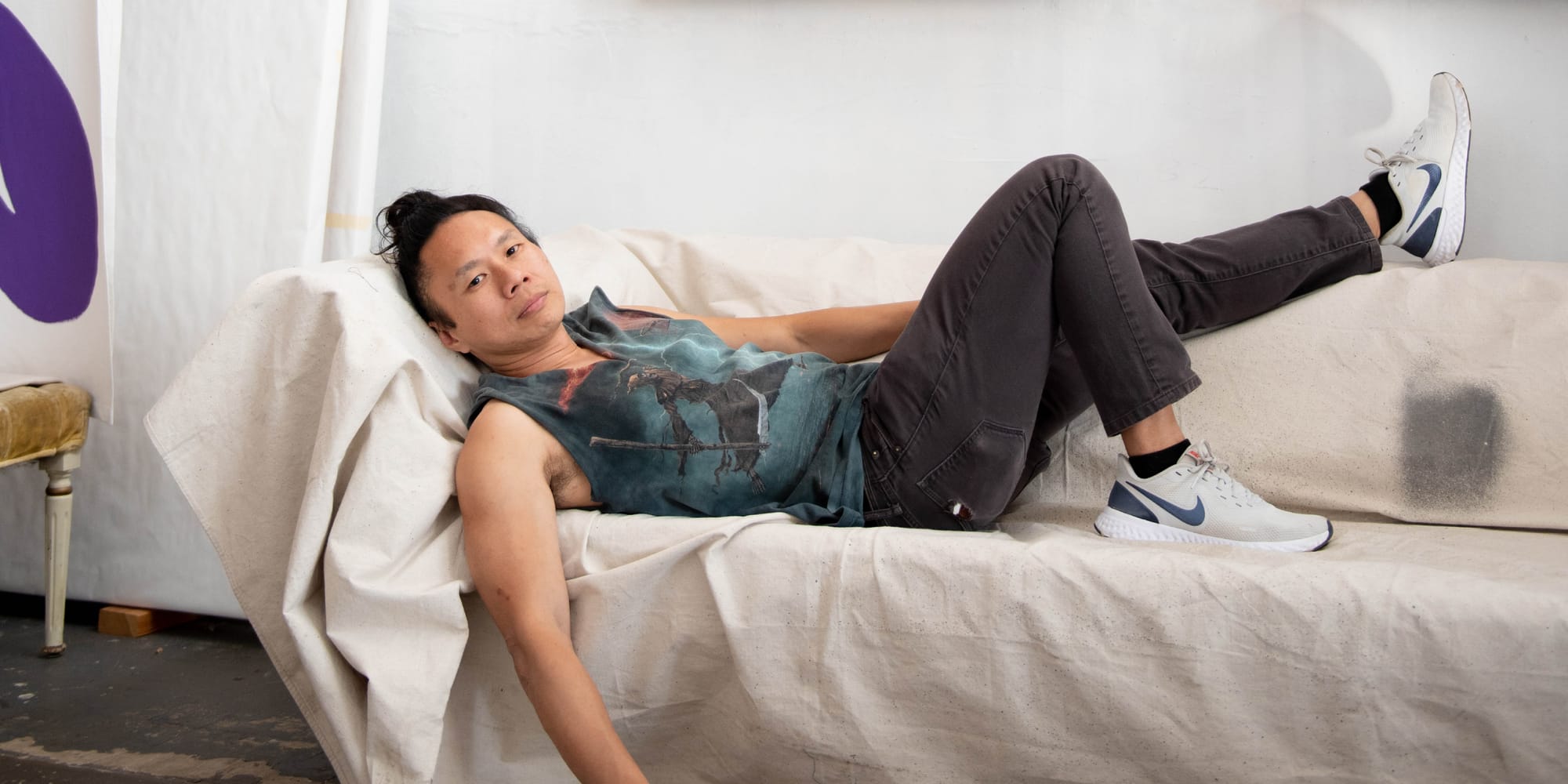 An individual dressed in a colorful tank top, dark jeans and white sneakers lays on a couch.