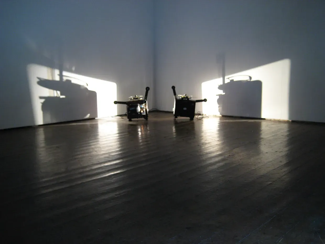 An empty corner of a gallery with two film projectors on the floor, with their large shadows cast onto the walls.