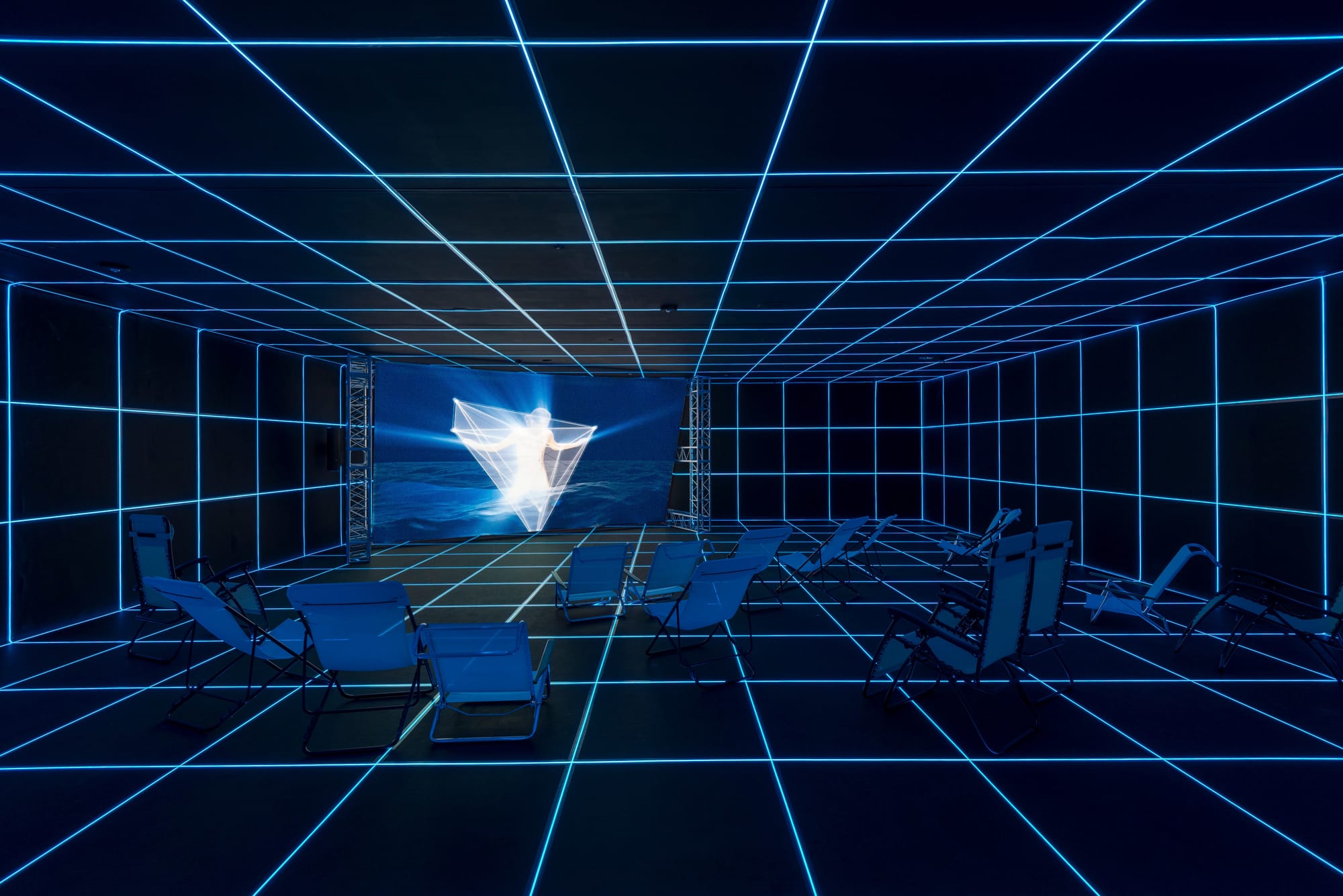 A dark gallery space designed to appear as an abstract digital grid, with blue lounge chairs positioned in front of a large video screen.