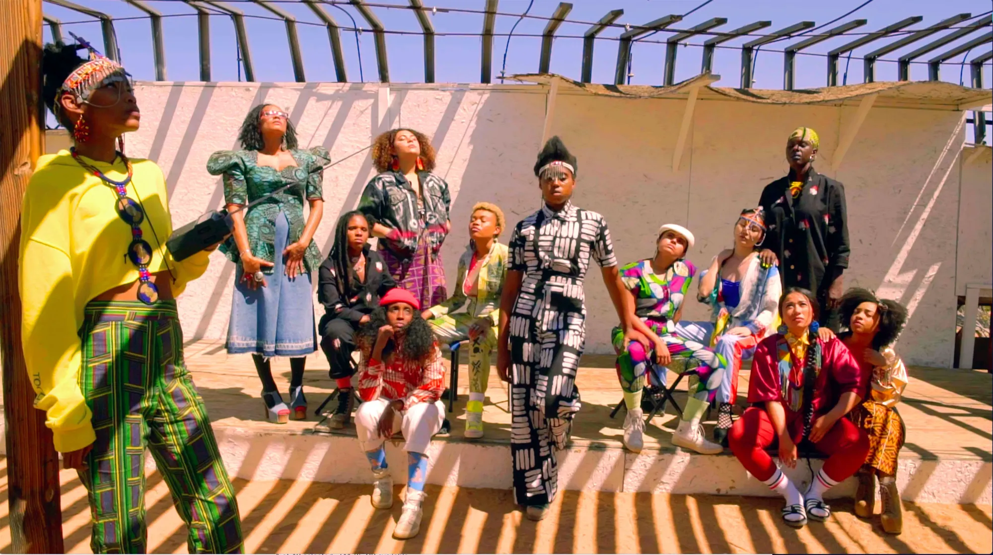 A group of Black women wearing colorful outfits in a variety of styles, posing in front of a tarp with slit shadows falling over them.
