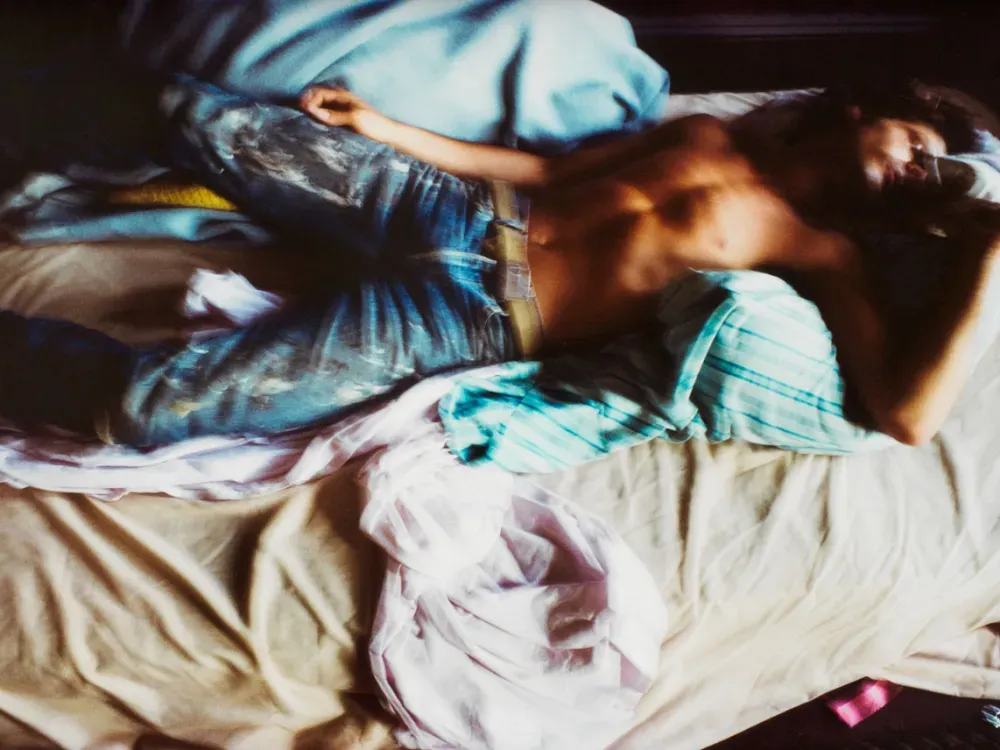 A blurry image of a shirtless man wearing jeans laying in a disheveled bed.