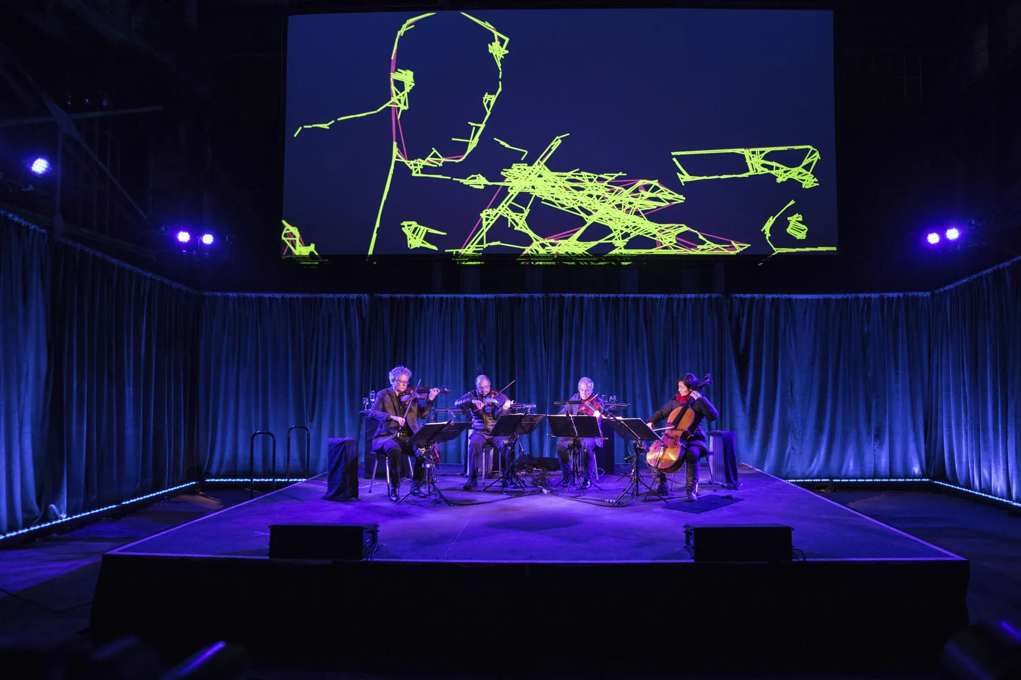 Four musicians on a dimly-lit stage perfom with string instruments with digital images projected on a screen behind them.