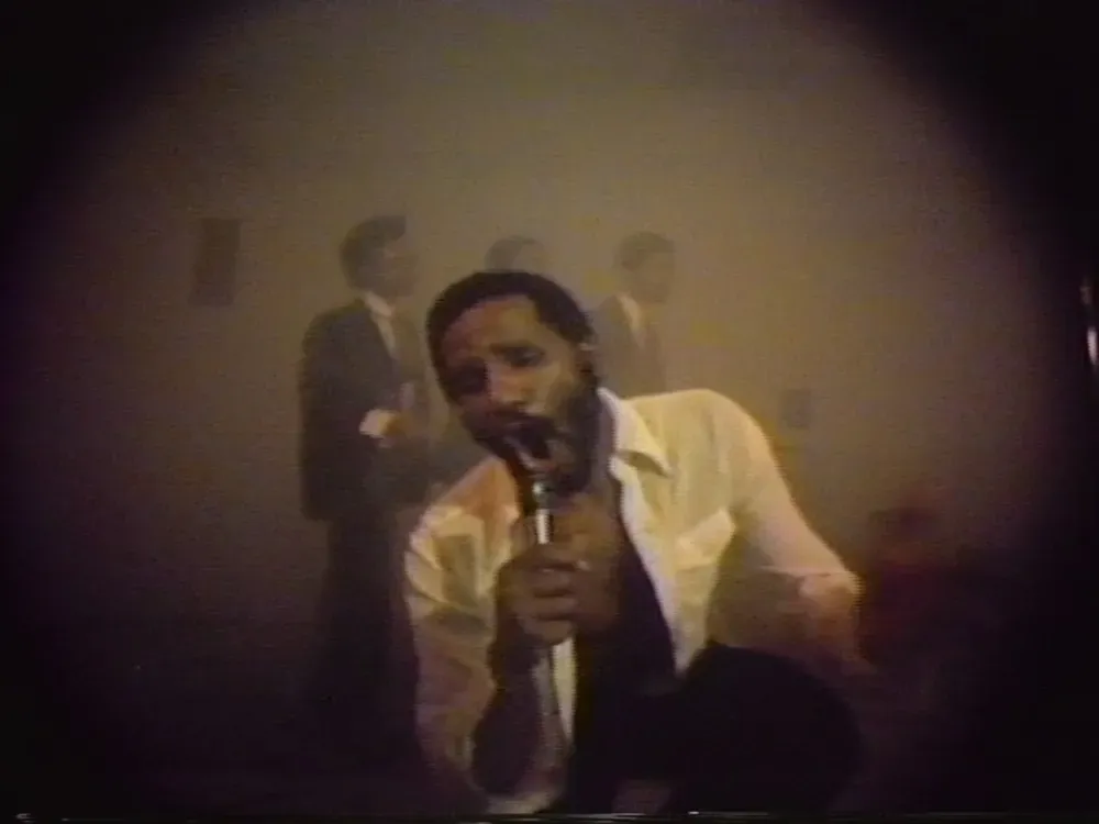 A blurry video image of a Black man singing into a microphone with his white shirt unbuttoned.