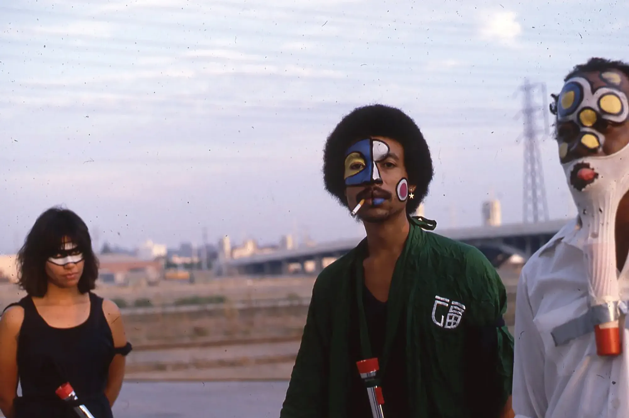 Three individuals with colorful face paint, wearing flashlights around their necks, standing against the Los Angeles city skyline in the background.