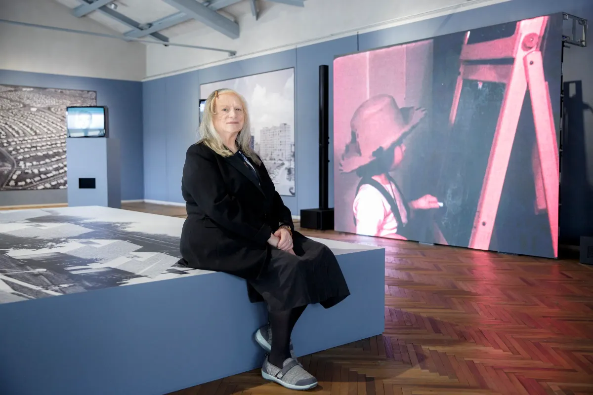 An older woman with a slight smile dressed in black sitting in the middle of a gallery exhibition.