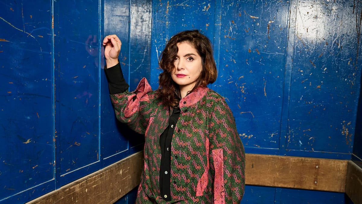 A woman with long dark brown hair, wearing a green and pink jacket, standing in a large service elevator painted blue.