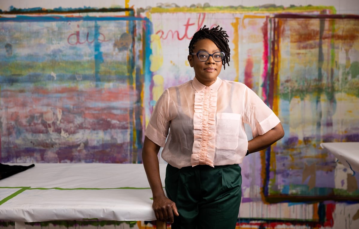 A Black woman with short dreadlocks and glasses, wearing a beige shirt and green pants, standing in front of a colorfully-painted wall.
