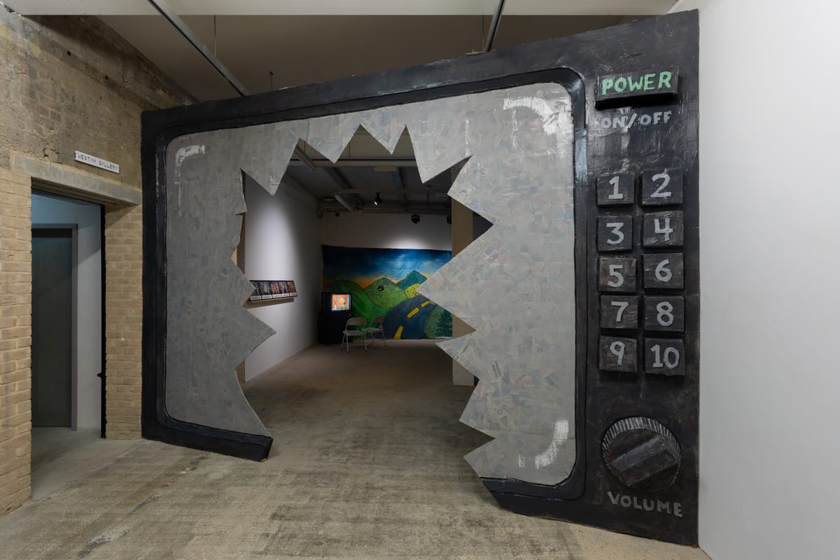A gallery view of an immense television with a broken screen that visitors walk through to access another gallery with monitors and tapestries.