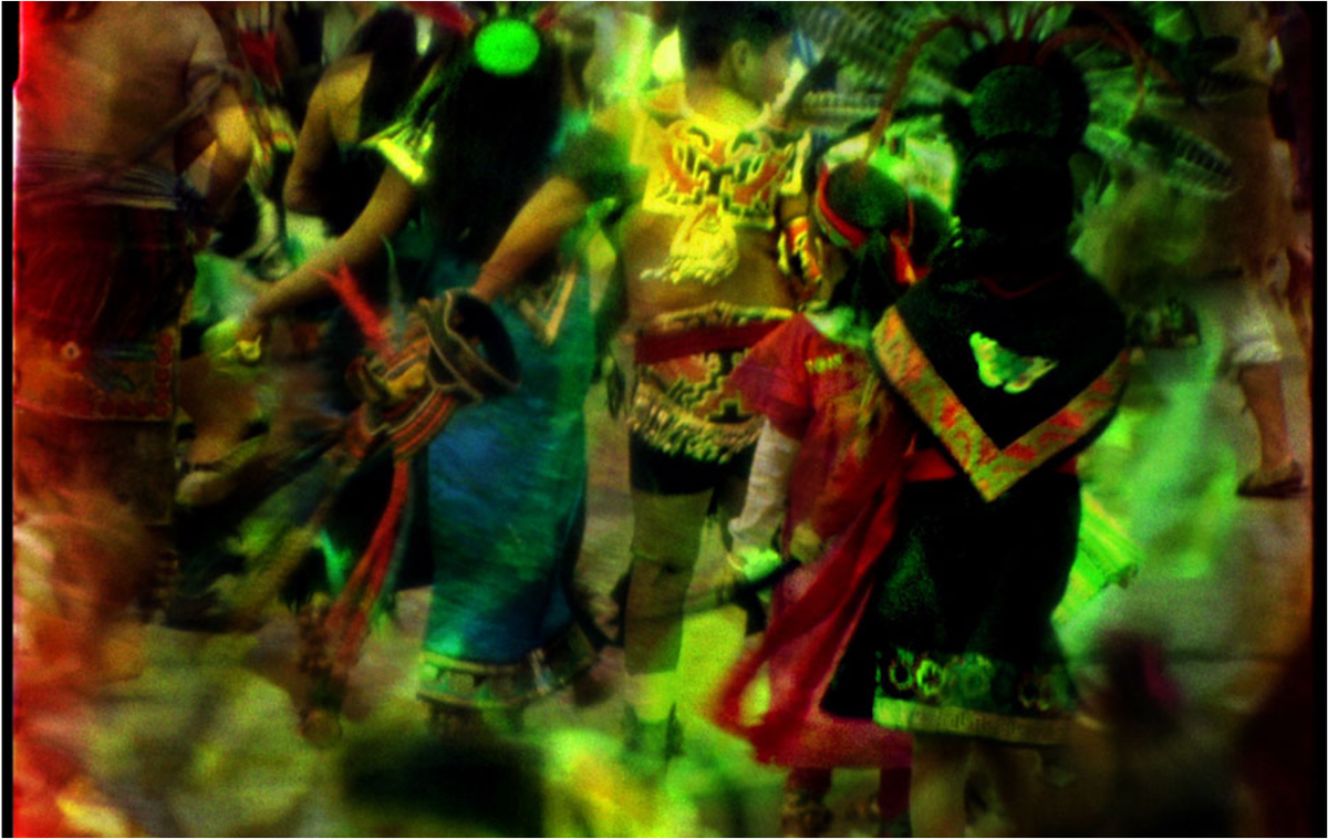A 16mm film frame with a lush, colorful view of Indigenous dancers in elaborate dress.