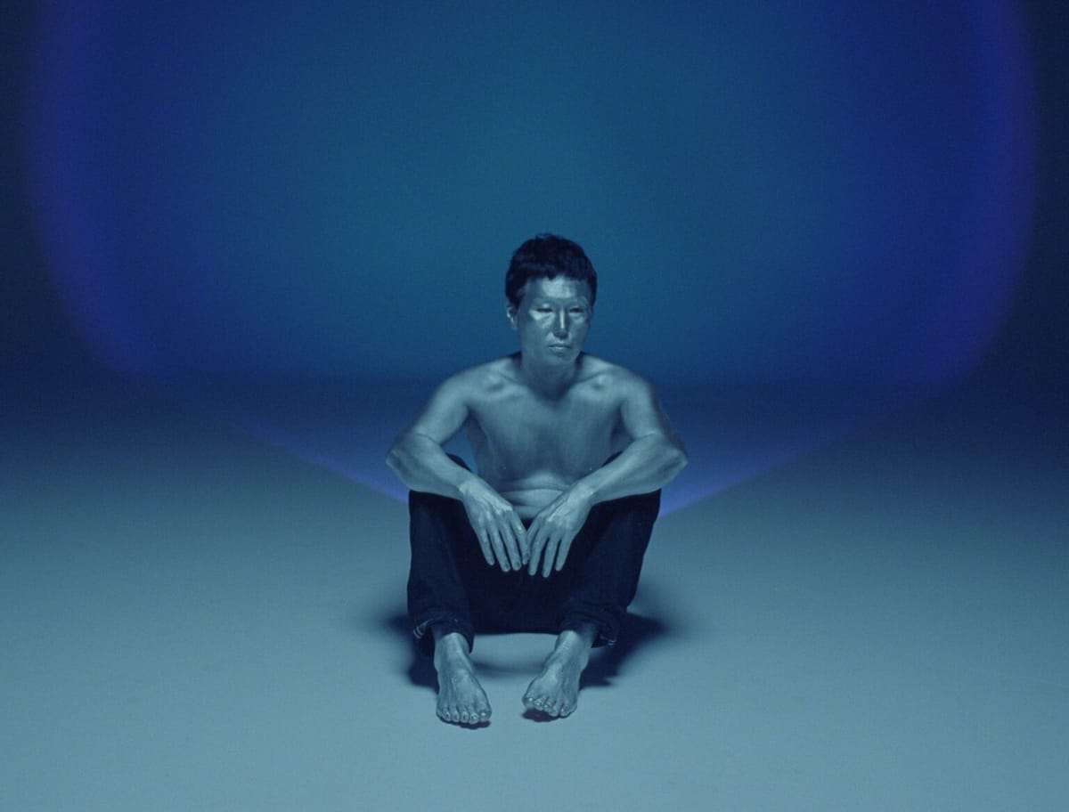 A man wearing black pants and no shirt sits against a nondescript turquoise backdrop, his gaze focused on something beyond the camera.