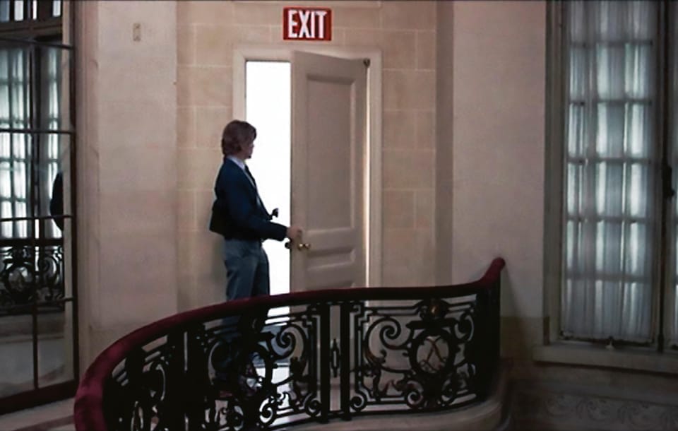 A man on an ornate balcony opening a door with an exit sign above it.