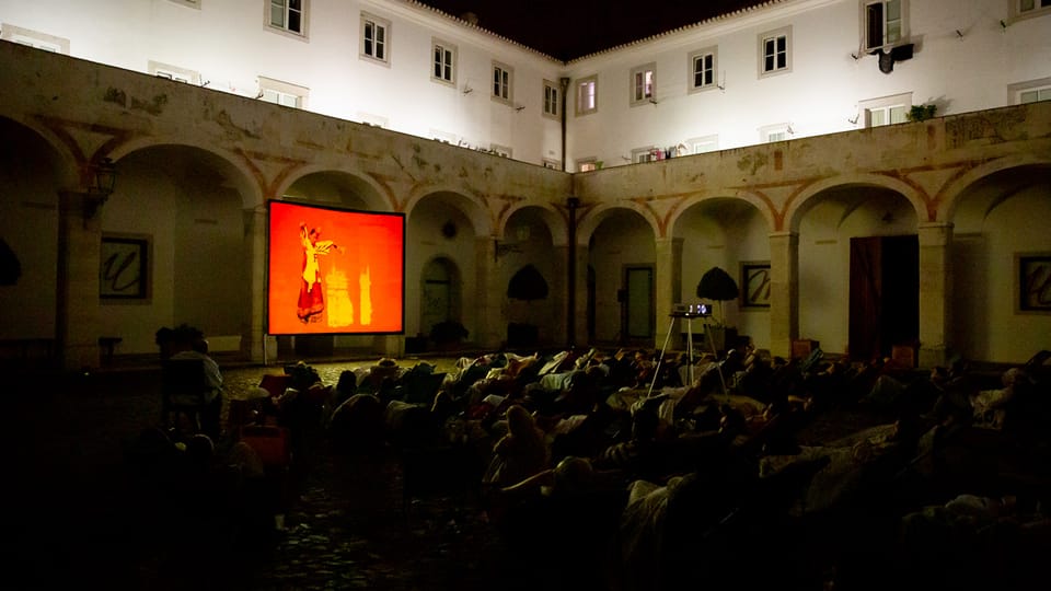 An open air screening for an audience in an archaic building, with a reddish-orange image of a dancer on screen.