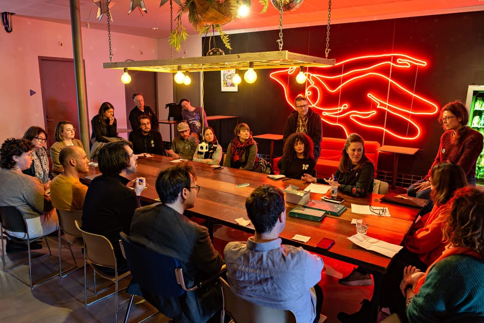 People at a conference sitting around a wooden table discussing with warm yellow light bulbs above them and a bright red neon sculpture on the wall behind them.