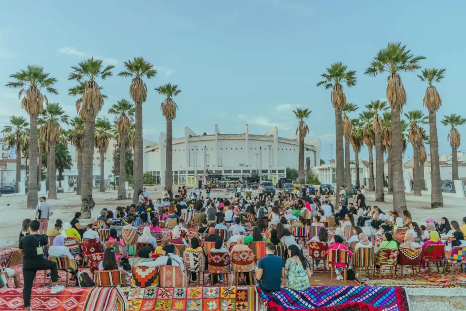 An open air gathering in a desert region with palm trees and audience members sitting on chairs with colorful patterns.