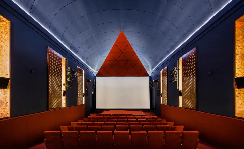 The interior of a cinema with a large red triangle above the screen, red plush seats, a blue ceiling, and golden wall panels.