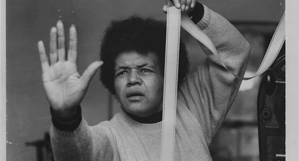 A black and white image of a Black woman holding a film strip in one hand, the other haid raised with palm up, and staring at something just off-camera.