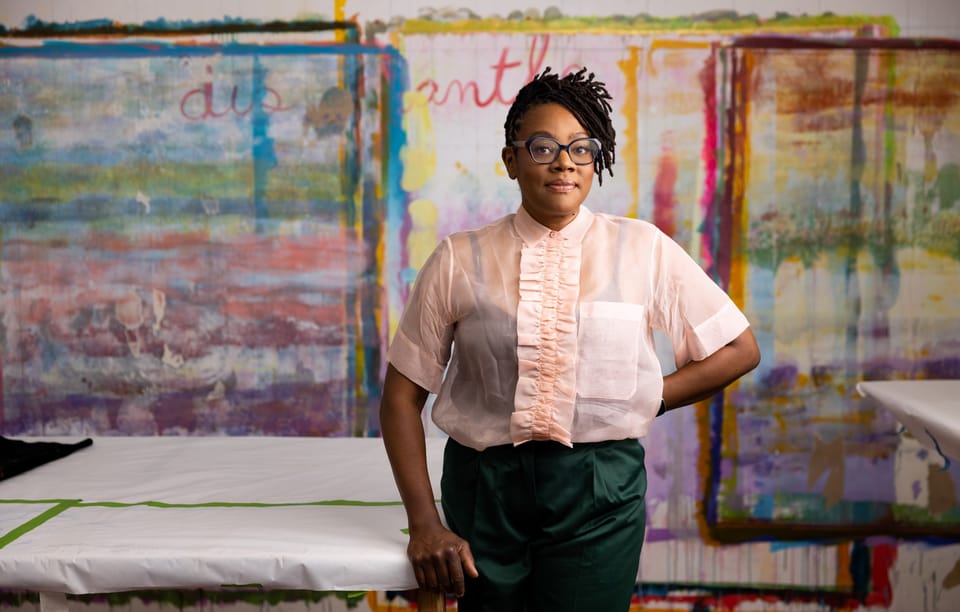 A Black woman with short dreadlocks and glasses, wearing a beige shirt and green pants, standing in front of a colorfully-painted wall.