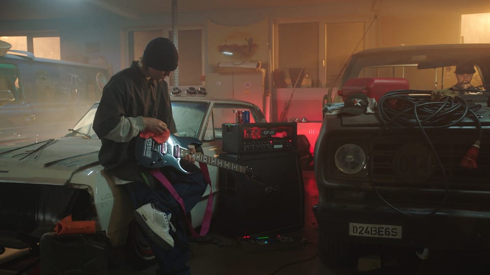 A hazily-lit garage with a man sitting on a car holding an electric guitar, with two other vehicles nearby in a state of disrepair