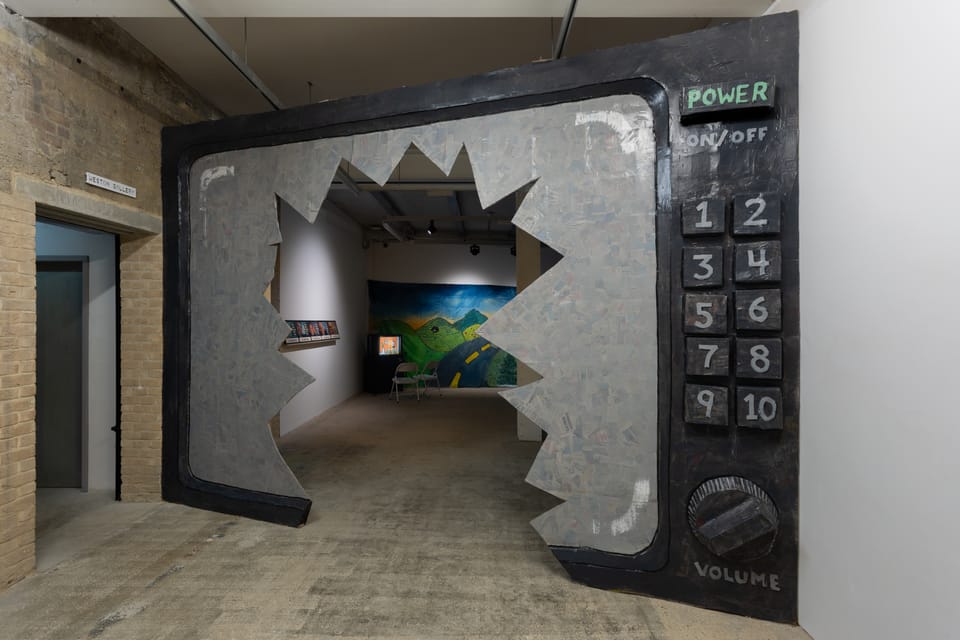 A gallery view of an immense television with a broken screen that visitors walk through to access another gallery with monitors and tapestries.