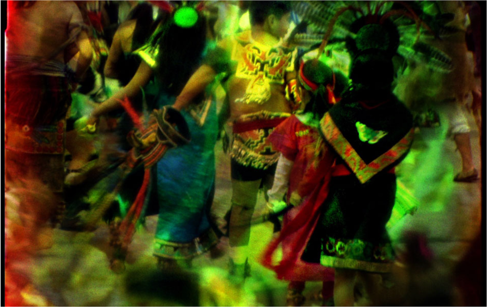 A 16mm film frame with a lush, colorful view of Indigenous dancers in elaborate dress.