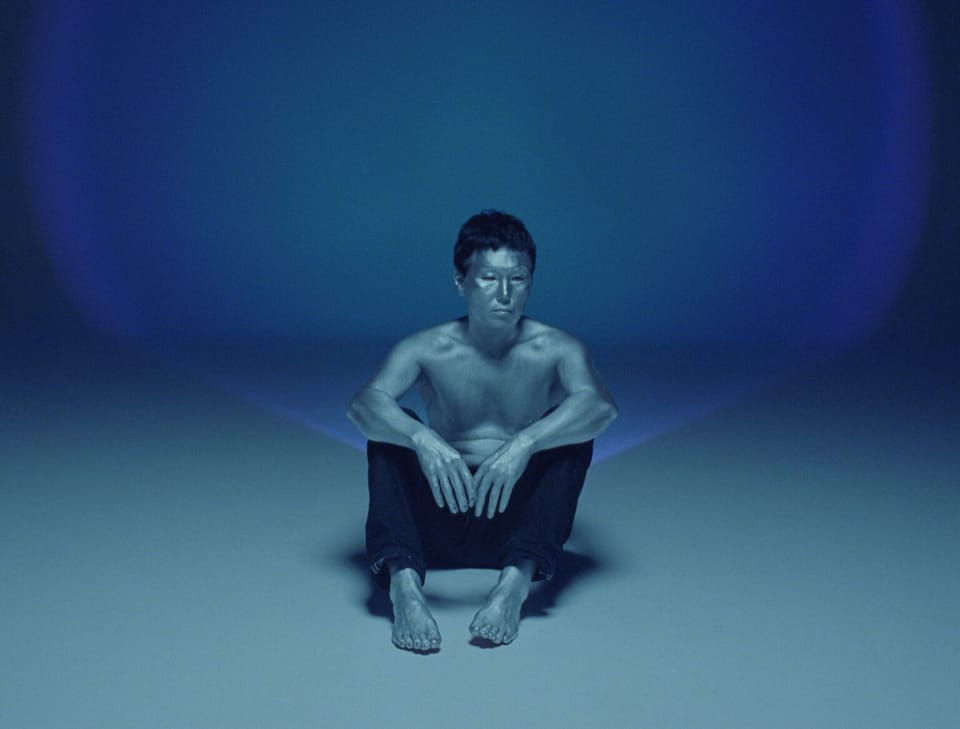 A man wearing black pants and no shirt sits against a nondescript turquoise backdrop, his gaze focused on something beyond the camera.